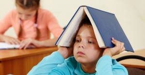 Cute schoolboy keeping open book on head in classroom